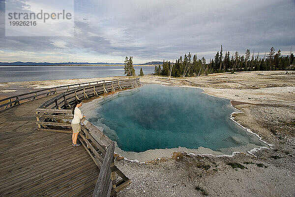 Eine erwachsene Frau steht auf der Promenade und blickt auf eine dampfende heiße Quelle am Yellowstone Lake im Yellowstone-Nationalpark  West