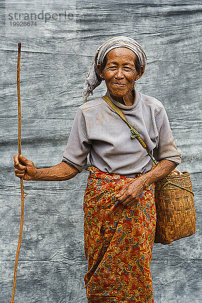 Porträt einer älteren Bäuerin  die mit Tasche und Zweig vor grauem Stoff posiert  Shan State  Myanmar