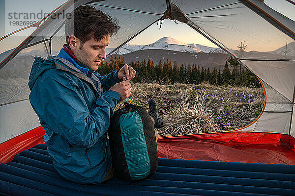Ein Wanderer packt Kleidung in einen Seesack  während er bei Sonnenuntergang auf seinem Zelt sitzt.