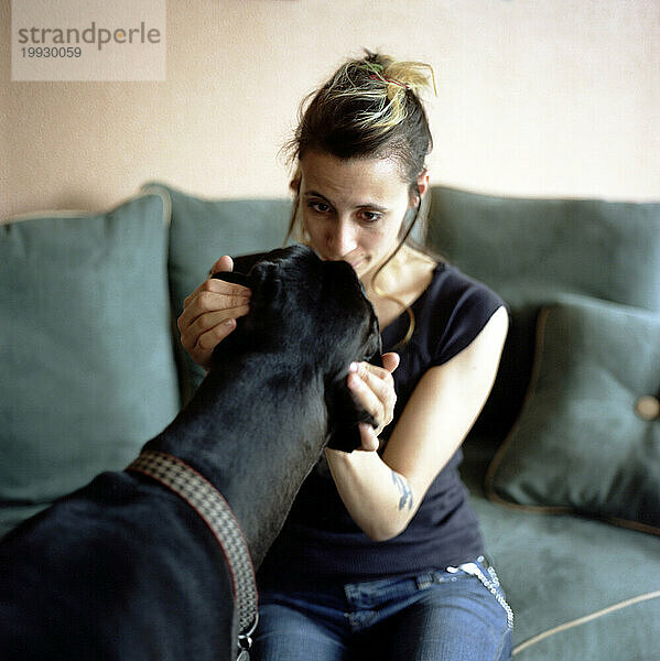 Junge Frau küsst den Scheitel ihres Hundes  während sie zu Hause auf der Couch sitzt (Deutsche Dogge).