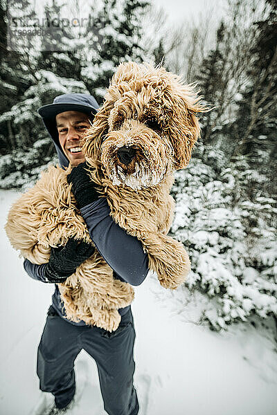 Lächelnder junger Mann hält seinen schneebedeckten Hund beim Spaziergang in New Hampshire