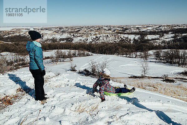 Weite Sicht auf Mädchen  die im malerischen Schnee auf dem Hügel rodeln
