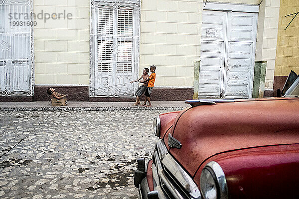 Drei Kinder spielen auf der Straße vor einem roten Oldtimer in Trinidad  Kuba