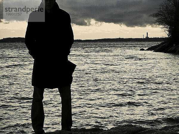 Ein Mann im Trenchcoat steht am Rande des Wassers  in der Ferne ein Fabrikschornstein. (Silhouette)