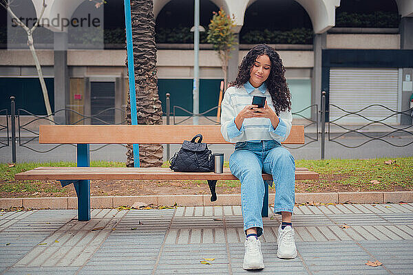 Junge erwachsene lateinamerikanische Frau mit lockigem Haar  die auf ihr Mobiltelefon schaut