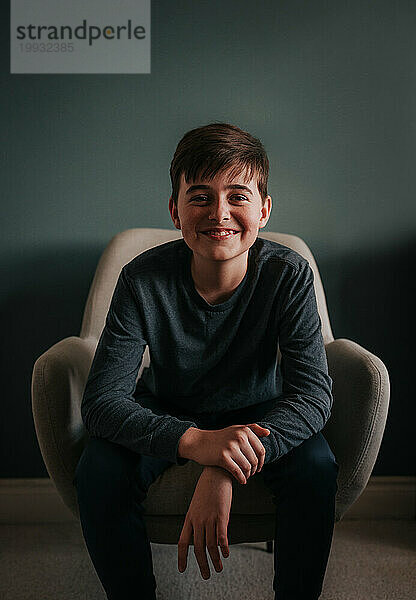 Porträt eines glücklichen Jungen  der drinnen auf einem Stuhl vor einer blauen Wand sitzt.