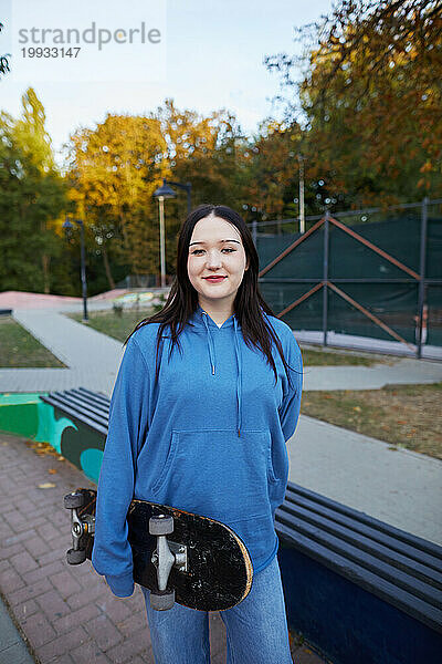 Lächelndes Mädchen  das mit einem Skateboard im Park steht