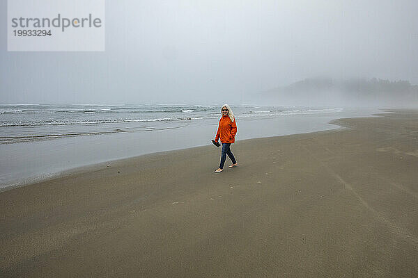 Frau in orangefarbener Jacke geht am nebligen Strand entlang