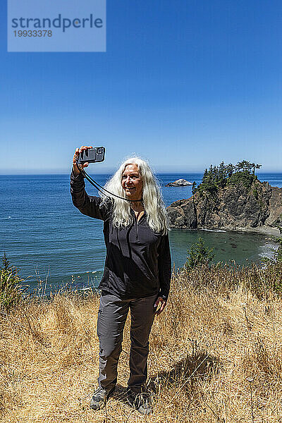 USA  Oregon  Brookings  Seniorin macht ein Selfie mit der Küstenlandschaft im Hintergrund