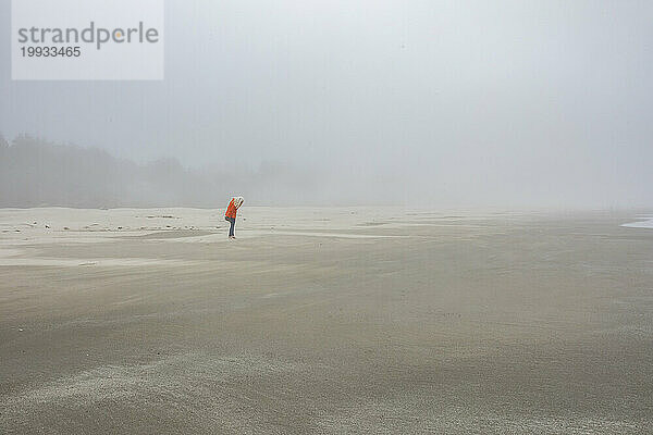 Frau in orangefarbener Jacke steht am nebligen Strand