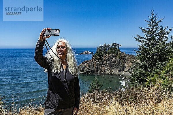USA  Oregon  Brookings  Seniorin macht ein Selfie mit Küstenlandschaft im Hintergrund