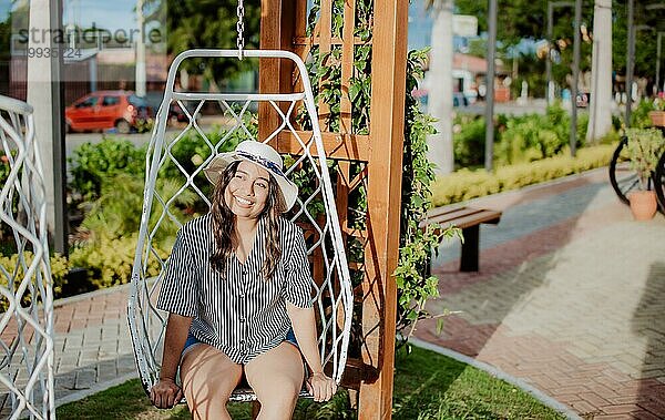 Ein lächelndes Mädchen sitzt auf einer Schaukel in La Calzada  Granada  Nicaragua. Entspanntes Mädchen mit Hut sitzt auf einer weißen Schaukel in einem schönen Garten