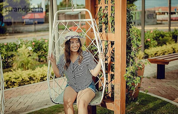 Entspanntes Mädchen mit Hut sitzt auf einer weißen Schaukel in einem schönen Garten. Ein lächelndes Mädchen sitzt auf einer Schaukel in La Calzada  Granada  Nicaragua  Mittelamerika