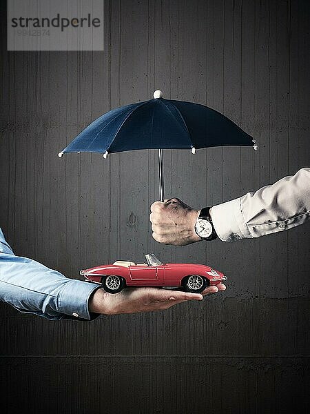 Geschäftsmann hält einen kleinen blaün Regenschirm zum Schutz eines Oldtimers  Schutz oder Autoversicherung Konzeptbild