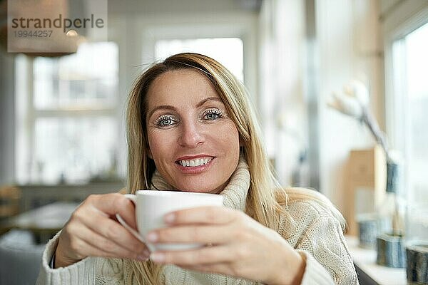 Porträt einer schönen Frau mit langen blonden Haaren  die lächelt und in die Kamera schaut  während sie sich zu Hause bei einer Tasse Kaffee entspannt