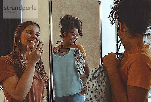 Zwei multiethnische Freundinnen haben Spaß beim Anprobieren von Kleidern vor dem Spiegel zu Hause. Ein lächelndes afroamerikanisches Mädchen hält ein Kleid auf einem Bügel und zeigt der daneben stehenden Freundin ihr Outfit. Rassenübergreifende Freundschaft
