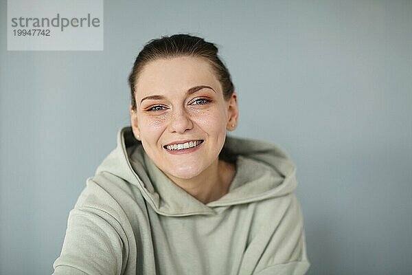 Wunderschönes Porträt einer jungen  fröhlichen 30er Jahre Frau in lässigem Sweatshirt  die gerade in die Kamera schaut und breit und glücklich lächelt und ihr glänzendes Glitzer Make up zeigt  posiert vor einem grauen Wandhintergrund