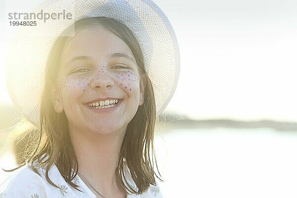 Fröhlicher verspielter weiblicher Teenager mit gemalten blaün Sommersprossen  der einen weißen Hut trägt und in die Kamera schaut  während er im Hintergrund am Strand in einem sonnigen Sommertag steht