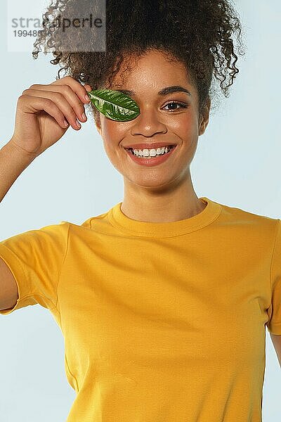 Natürliche Schönheit. Glückliches afroamerikanisches Mädchen in gelbem TShirt mit lockigem Haar  das ein Auge mit grünem Blatt bedeckt  breit lächelnd  während sie direkt in die Kamera schaut  posiert vor hellem Wandhintergrund