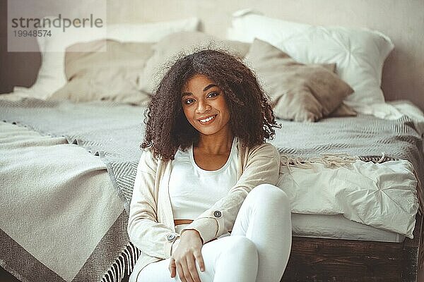 Fröhliche  charmante afroamerikanische junge Frau mit lockigem Haar im Schlafzimmer  sitzt auf dem Boden am Fußende des gemachten Doppelbetts  schaut mit breitem Lächeln in die Kamera  zufriedene Afro Frau  die sich zu Hause entspannt