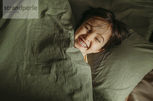 Glücklicher Junge  der zu Hause unter einer Decke im Bett liegt