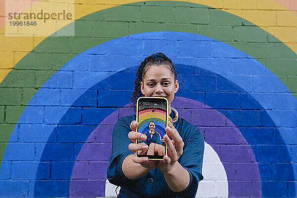 Lächelnde junge Frau zeigt Foto auf Smartphone vor Regenbogenwand