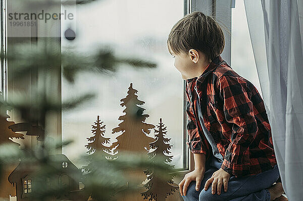 Boy near Christmas decoration looking through window at home