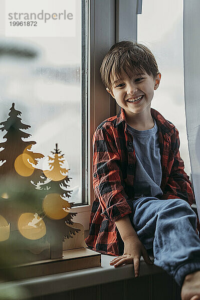 Lächelnder Junge sitzt auf der Fensterbank neben der Weihnachtsdekoration zu Hause