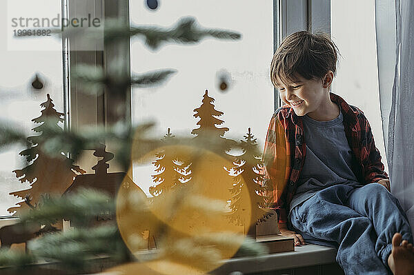Lächelnder Junge bewundert Weihnachtsdekoration am Fenster zu Hause