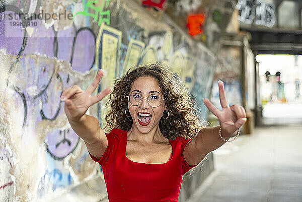 Fröhliche Frau gestikuliert Friedenszeichen in der Nähe der Graffitiwand auf dem Fußweg