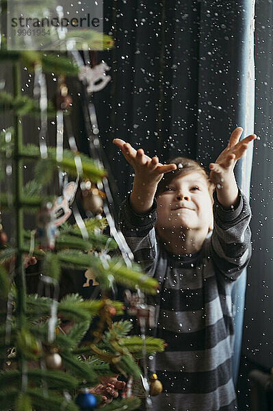 Verspielter Junge fängt zu Hause Kunstschnee neben dem Weihnachtsbaum