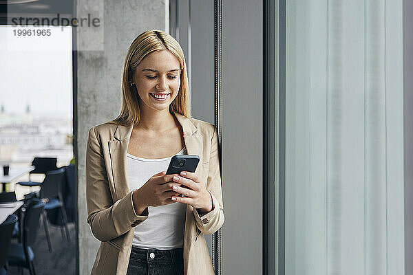 Glückliche Geschäftsfrau benutzt Smartphone am Fenster im Büro