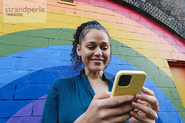 Lächelnde junge Frau benutzt Smartphone vor Regenbogenwand