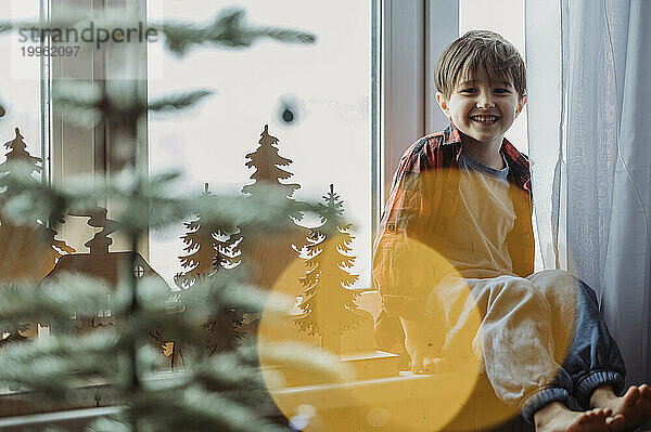 Smiling boy sitting on window sill near Christmas decoration at home