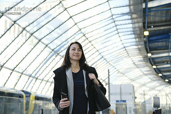 Lächelnde reife Frau mit Handtasche und Smartphone am Bahnhof