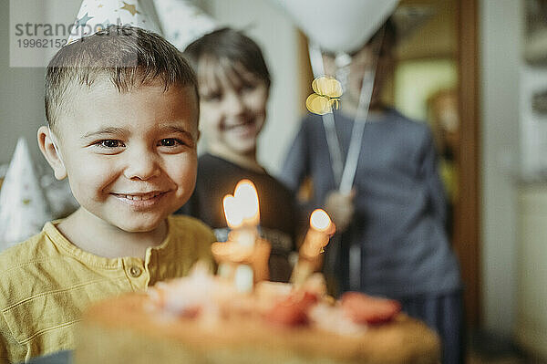 Fröhlicher Junge mit Geburtstagstorte und Geschwistern im Hintergrund