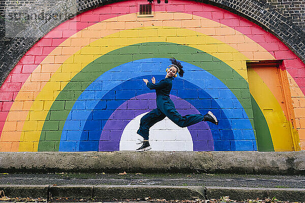 Fröhliche junge Frau trägt kabellose Kopfhörer und tanzt vor der Regenbogenwand