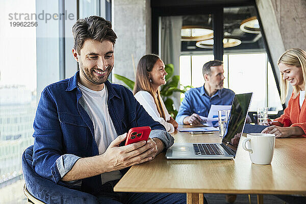 Glücklicher junger Geschäftsmann  der sein Smartphone in der Nähe von Kollegen im Büro nutzt