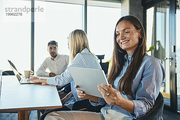 Glückliche Geschäftsfrau nutzt Tablet-PC mit Kollegen  die im Hintergrund diskutieren