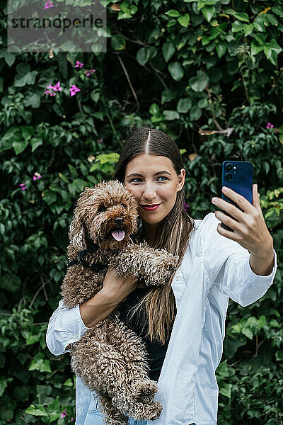 Lächelnde Frau macht Selfie mit Pudelhund vor Pflanzen