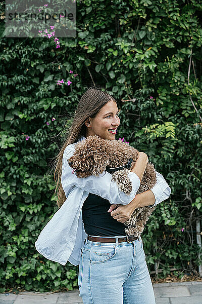 Lächelnde junge Frau hält Pudelhund in der Hand und steht vor Pflanzen