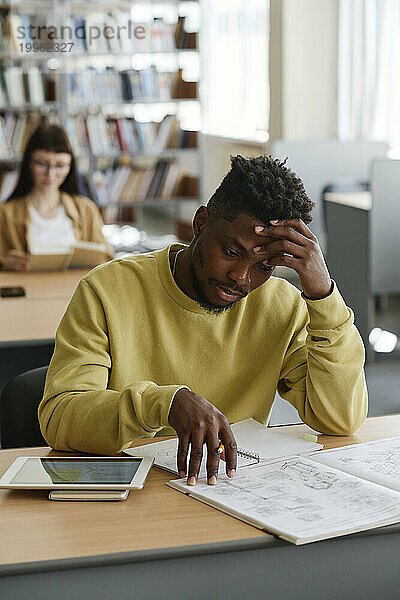 Müder Student mit Kopf in der Hand liest Buch in der Bibliothek