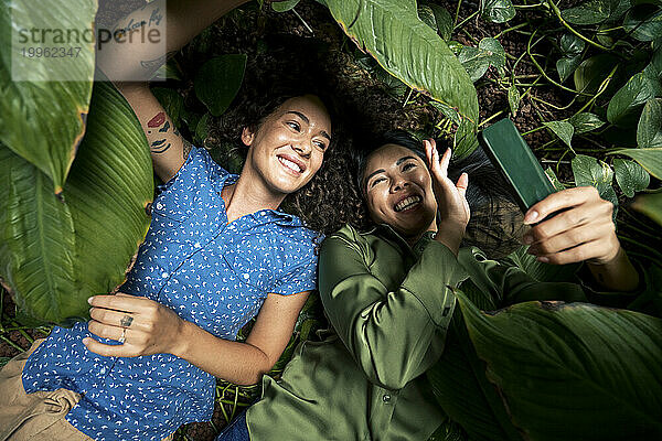 Glückliche Frau  die mit einer Freundin inmitten von Pflanzen ein Selfie mit dem Smartphone macht