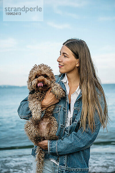 Lächelnde junge Frau mit Pudelhund steht am Strand