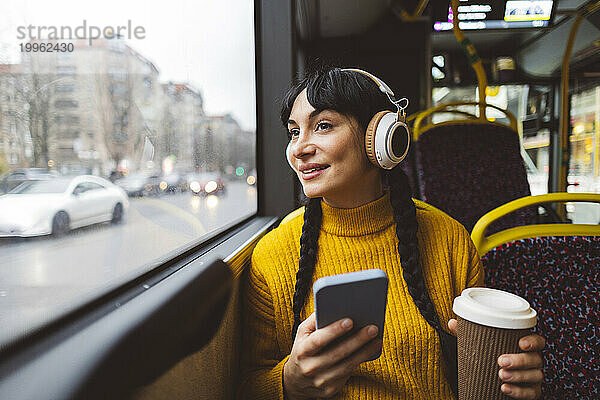 Glückliche Frau trägt kabellose Kopfhörer und hält Kaffeetasse mit Smartphone im Bus