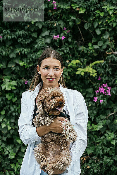 Lächelnde junge Frau mit Pudelhund steht vor Pflanzen