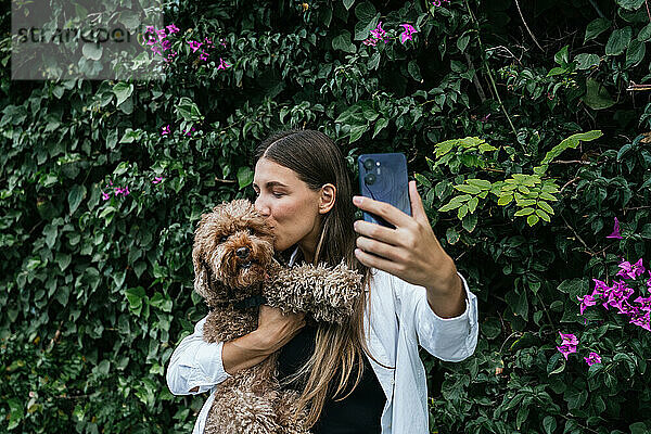 Junge Frau küsst und macht Selfie mit Pudelhund vor Pflanzen