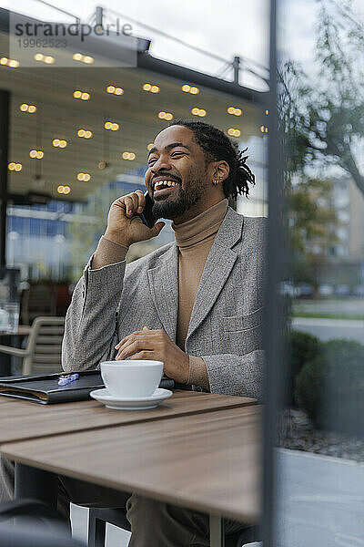 Glücklicher Geschäftsmann  der im Büropark mit Kaffee sitzt und mit dem Smartphone spricht