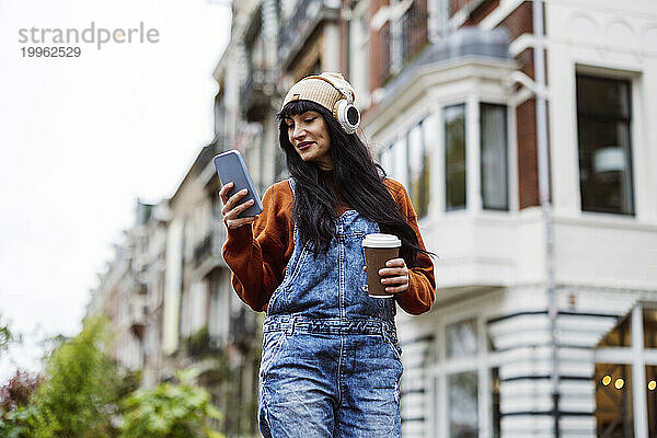 Lächelnde schöne Frau hält Kaffeetasse in der Hand und benutzt Smartphone vor Gebäuden