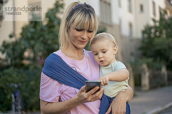 Sohn im Tragetuch benutzt Smartphone  gehalten von Mutter  die auf der Straße steht